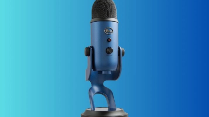 Setup desk minimalis dengan microphone Blue Yeti silver terpasang pada boom arm di samping monitor komputer yang menampilkan software editing audio.