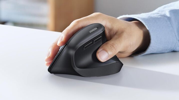 Mouse vertikal UGREEN hitam dengan desain ergonomis diletakkan di atas meja kayu minimalis di samping laptop modern.