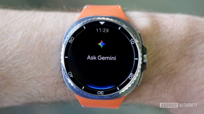 Tampilan layar smartwatch Wear OS menunjukkan antarmuka cuaca Gemini terbaru dengan ikon grid interaktif di atas meja kayu